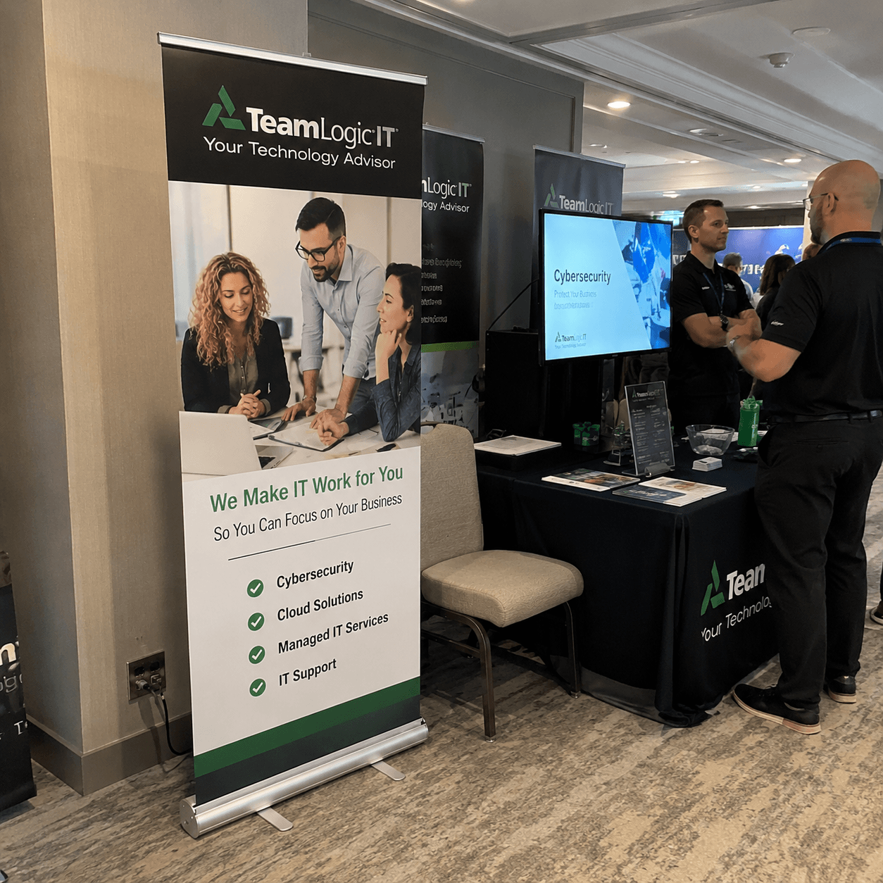 TeamLogic IT booth at a conference with a large retractable banner and a screen displaying cybersecurity information.
